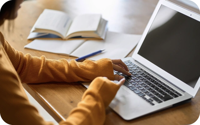 Sieviete dzeltenā džemperī raksta portatīvajā datorā, kas novietots uz koka galda; blakus atvērta grāmata un pierakstu klade ar pildspalvu.