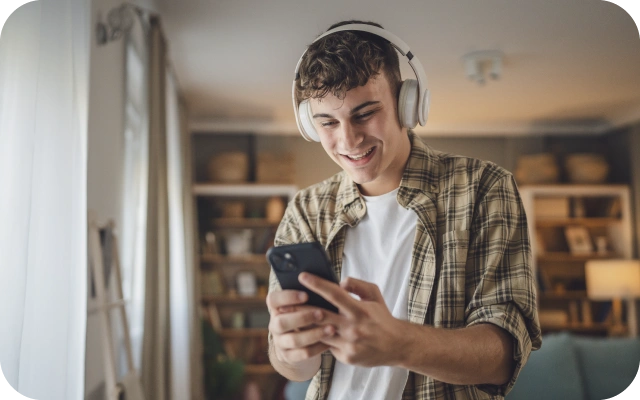 Jaunietis rūtainā kreklā un baltā T-kreklā ar lielām austiņām ap galvu, stāv telpā ar grāmatu plauktiem fonā, turot rokās mobilo telefonu un skatoties tajā.