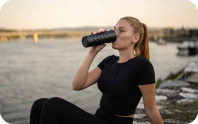 Sieviete melnā sporta tērpā sēž pie ūdens tilpnes un dzer no melnas pudeles.