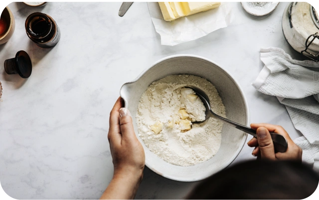 Bļodā uz galda tiek sajaukoti milti un sviesta gabaliņi ar karoti, fonā sastāvdaļas — sviesta paku, trauks ar miltiem, mazās pudelītes un virtuves dvielis uz gaišas virsmas.