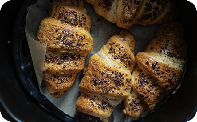 Gaisa fritera grozā novietoti svaigi cepti kruasāni ar zeltaini brūnu garoziņu un šokolādes gabaliņu pārklājumu, uz cepampapīra karstā gaisa fritera pamatnes.
