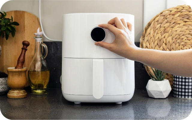 Balts gaisa friteris novietots uz virtuves virsmas, persona pielāgo temperatūras vai laika regulatoru; fonā virtuves piederumi, eļļas pudele un pinuma paliktnis.