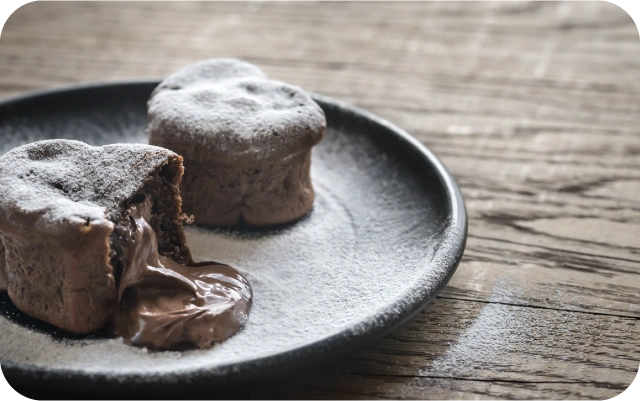Divi šokolādes lava kēksi uz melnas šķīvja malas, viens pārgriezts ar izplūstošu šokolādes pildījumu, deserts apkaisīts ar pūdercukuru uz koka galda virsmas.