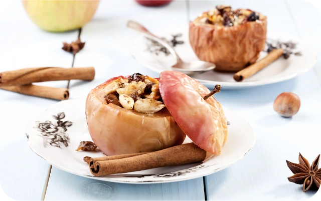 Cepti pildīti āboli ar riekstiem un rozīnēm pasniegti uz baltiem dekoratīviem šķīvjiem, blakus kanēļa standziņas, zvaigžņu anīss un lazdurieksts uz gaiša koka galda.