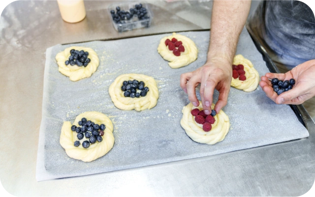 Uz cepampapīra novietotas apaļas rauga mīklas masas ar iedobēm, kurās tiek liktas mellenes un avenes; redzamas rokas, kas uzliek ogas, fonā nerūsējošā tērauda darba virsma un ogu trauciņš.