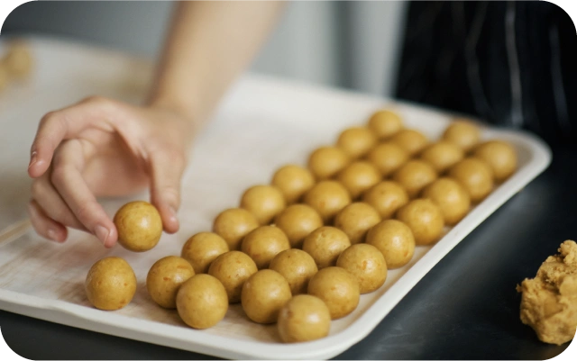 Uz paplātes izkārtotas vienāda izmēra mīklas bumbiņas, blakus rokas, kas novieto vienu no bumbiņām, fonā atrodas neliels mīklas gabals uz darba virsmas.