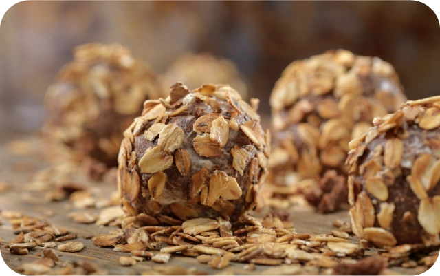 Tuvplānā apaļas bumbiņas, kas pārklātas ar auzu pārslām un izvietotas uz koka virsmas ar izbirušām pārslām apkārt.