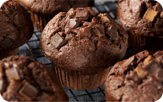 Tuvplāns ar vairākiem šokolādes mafiniem un šokolādes gabaliņiem uz tiem, mafini novietoti uz dzesēšanas restes.