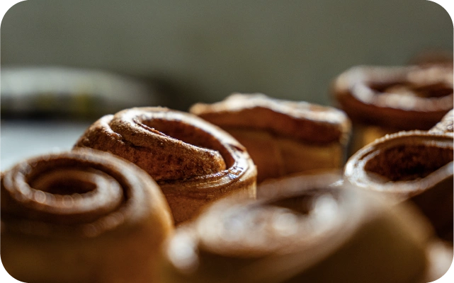 Tuvu nofotografētas svaigi ceptas kanēļmaizītes ar redzamu spirāles tekstūru un zeltaini brūnu virsmu uz neitrāla fona.