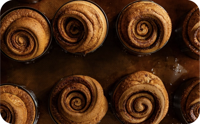 No augšas fotografētas svaigi ceptas kanēļmaizītes ar spirālveida pārritinātu mīklu, izkārtotas vairākās rindās uz cepampannas ar brūnu cepampapīru.