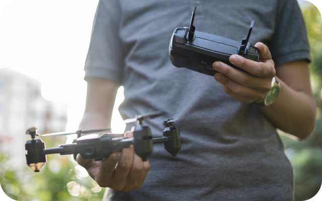 Vīrietis pelēkā kreklā tur rokās dronu un tālvadības pulti, gatavojoties pacelt dronu gaisā.