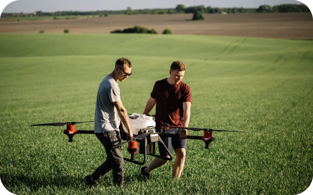 Divi vīrieši tumšā apģērbā pa labības laukā nes lielu dronu ar četriem propelleriem.