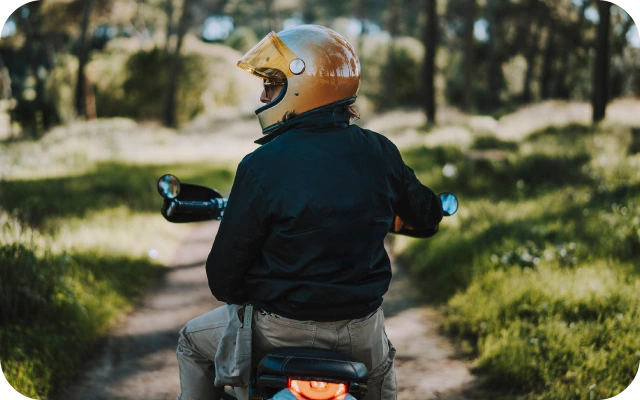 Persona ar dzeltenu motocikla ķiveri sēž uz motorollera un brauc pa šauru meža taku, fonā zaļa daba un koki.