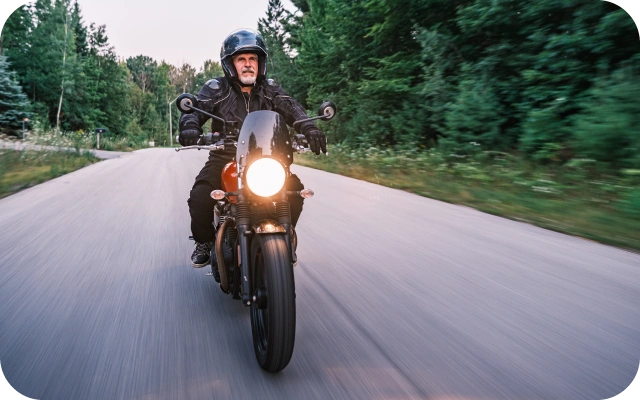 Motocikls ar ieslēgtu priekšējo lukturi brauc pa asfaltētu ceļu mežainā apvidū, braucējs valkā aizsargķiveri un ekipējumu, fonā koki un izplūdusi kustības perspektīva.