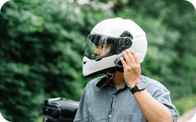 Vīrietis ar baltu motocikla ķiveri pieskaras ķiveres sānu daļai, regulējot intercom sakariem; vīrietim uz rokas pulkstenis, fonā zaļš, dabīgs apvidus ar kokiem.
