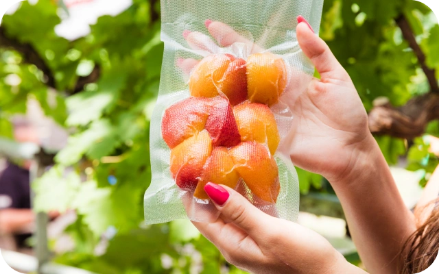 Sieviete rokās tur vakuumā iepakotus persikus, fonā zaļas koku lapas.