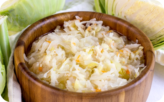 Koka bļoda ar skābētiem kāpostiem uz galda, fonā svaigi kāposti.