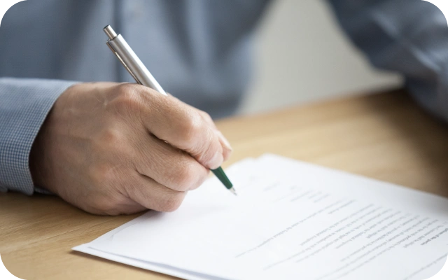 Vīrietis zilā kreklā raksta ar pildspalvu uz dokumenta, kas novietots uz koka galda.