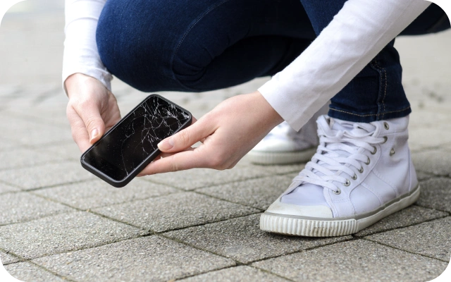 Persona baltās kedās un zilos džinsos tup uz bruģēta ceļa, turot mobilo telefonu ar saplīsušu ekrānu.