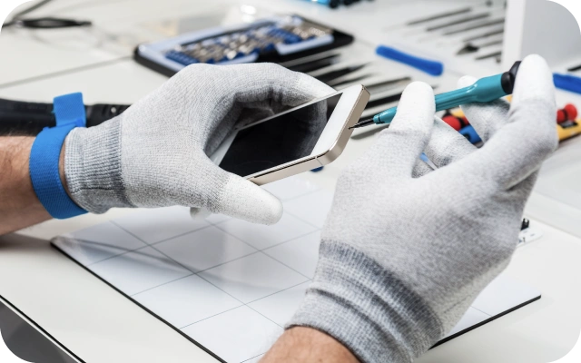 Rokas pelēkos cimdos veic mobilā telefona remontu, izmantojot skrūvgriezi; fonā darba galds ar dažādiem instrumentiem un piederumiem.