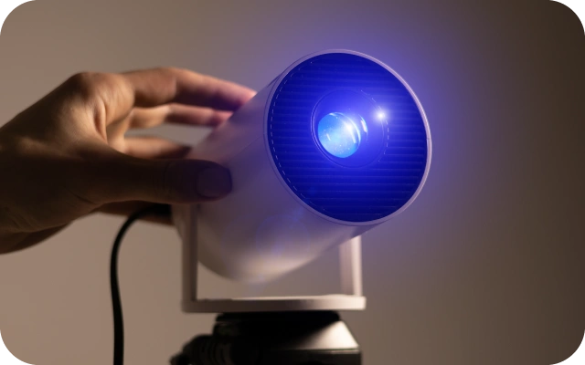 Rokas pielāgo balta kompaktā projektora pozīciju, ierīcei spīdot ar intensīvu zilu gaismu tumšā telpā.