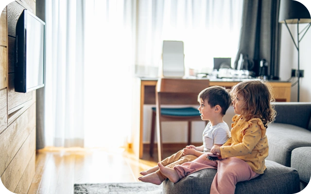 Divi mazi bērni sēž uz dīvāna gaišā dzīvojamajā istabā un skatās televizoru, viens bērns tur pulti; fonā ir galds, krēsls un liels logs ar aizkariem.