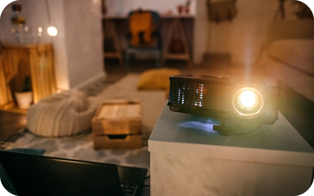 Ieslēgts projektors uz balta galda mājīgā istabā ar siltu apgaismojumu, kur fonā ir mēbeles, paklājs un spilveni.