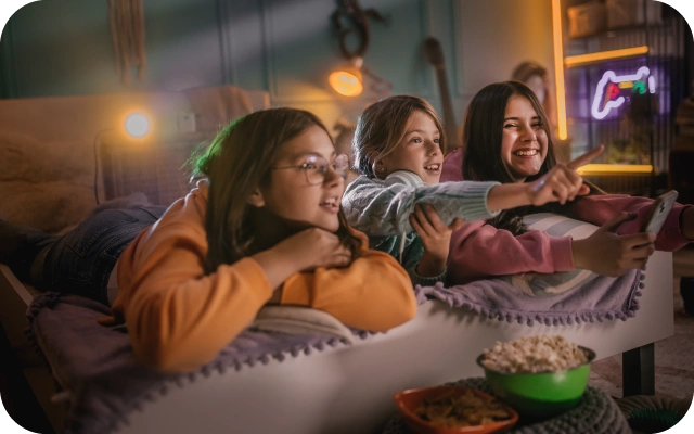 Trīs meitenes atrodas viesistabā, guļot uz dīvāna un skatoties ekrāna virzienā, vienai meitenei rokā ir mobilais telefons; priekšplānā redzama bļoda ar popkornu un citām uzkodām. Telpas aizmugurē spīd spoža gaisma no projektora.