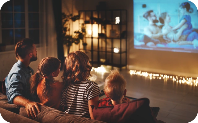 Ģimene sēž uz dīvāna mājīgā viesistabā un skatās sienas projekciju ar filmu, telpā redzamas dekoratīvas lampiņas un plaukts ar apgaismojumu.