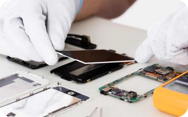 Viedtālruņa remonts uz darba galda – baltas, cimdotas rokas noņem telefona displeju, apkārt izkārtotas ierīces detaļas, piemēram, mātesplate, akumulators un korpusa elementi; instrumenti un elektronikas komponentes uz gaišas virsmas.