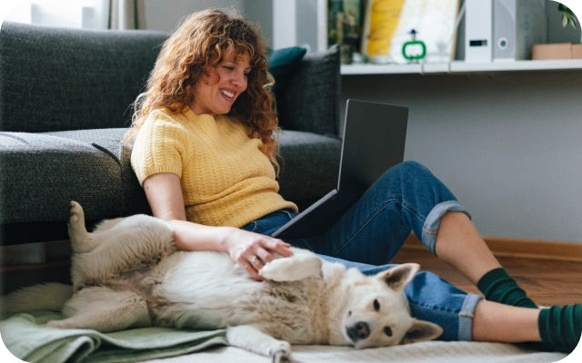 Sieviete sēž uz grīdas pie dīvāna mājas interjerā un strādā ar portatīvo datoru, blakus guļ suns uz segas; fonā plaukti un interjera dekori, telpā dabīgs apgaismojums.