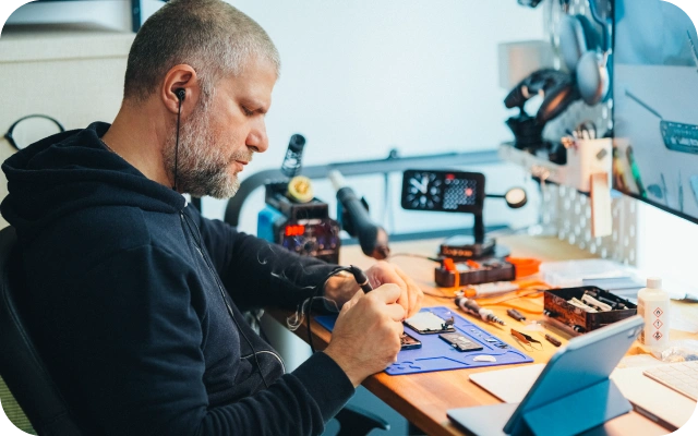 Vīrietis darbnīcā sēž pie darba galda un veic elektronikas remontu, strādājot ar nelielām detaļām uz antistatiskas paklāja; uz galda lodēšanas instrumenti, skrūvgrieži, elektronikas komponentes, diagnostikas iekārtas, monitors un planšetdators, fonā tehnikas piederumi un darba apgaismojums.