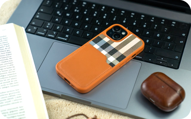 Viedtālrunis ar oranžu ādas tipa aizsargvāciņu un rūtainu dizaina paneli novietots uz portatīvā datora tastatūras; blakus bezvadu austiņu uzlādes kārbiņa un atvērta grāmata uz gaišas virsmas, veidojot darba un ikdienas piederumu kompozīciju.