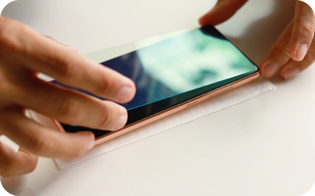 Tuvplāna skats uz viedtālruni uz balta galda, uz kura ar rokām tiek uzklāts ekrāna aizsargstikls vai aizsargplēve; precīza stikla izlīdzināšana uz telefona displeja, uzsverot viedtālruņa ekrāna aizsardzības procesu.