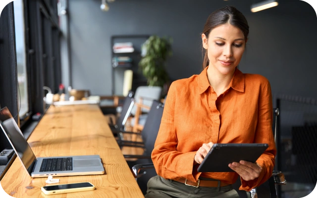 Sieviete birojā sēž pie gara koka galda un izmanto planšetdatoru; uz galda atvērts portatīvais dators, mobilais telefons un piederumi, fonā moderns darba interjers ar krēsliem un augiem, telpā dabīgs apgaismojums no loga.