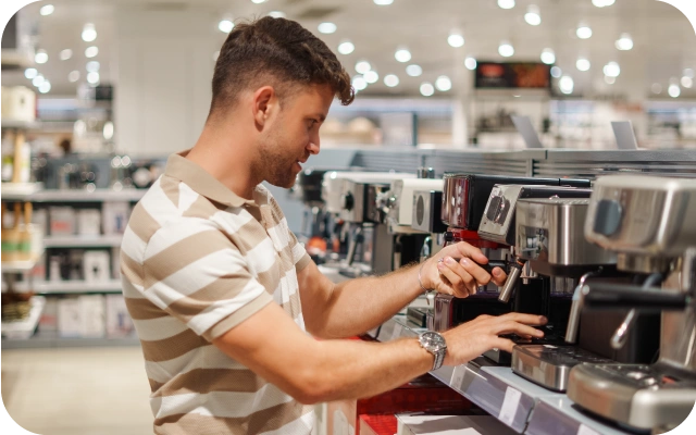 Vīrietis elektropreču veikalā pārbauda espresso kafijas automātu, pagriežot tā tvaika vai ūdens padeves sviru, fonā kafijas automātu rindas plauktos.