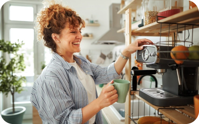 Sieviete virtuvē pie plaukta darbojas ar kafijas automātu, turot krūzi vienā rokā, bet ar otru roku pagriežot ierīces vadības pogu.
