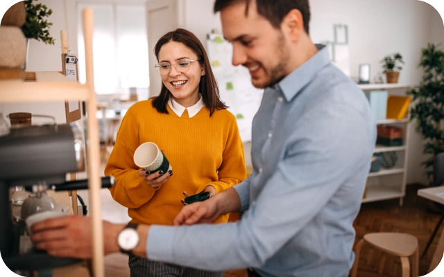 Divi cilvēki virtuvē darbojas ar kafijas automātu; sieviete tur krūzi rokā, bet vīrietis savu krūzi novieto zem kafijas automāta iztekas, fonā plaukti un virtuves piederumi.