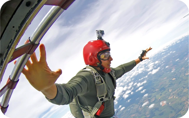 Vīrietis izlec no lidmašīnas ar izpletni; viņam galvā sarkana aizsargķivere ar piestiprinātu kameru un pelēku izpletņa sistēmu. Fonā ainava ar mākoņiem un zemes reljefu.