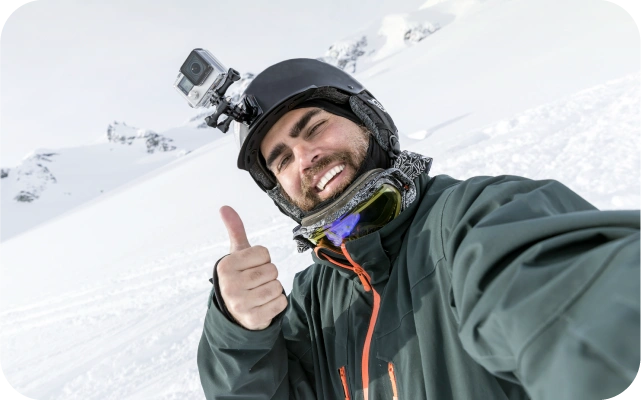 Vīrietis ziemas apģērbā ar melnu aizsargķiveri galvā, stāv sniegotā kalnu ainavā un uzņem pašbildi, rādot paceltu īkšķi. Pie ķiveres piestiprināta kamera. Fonā sniegoti kalnu virsotņu nogāzes.