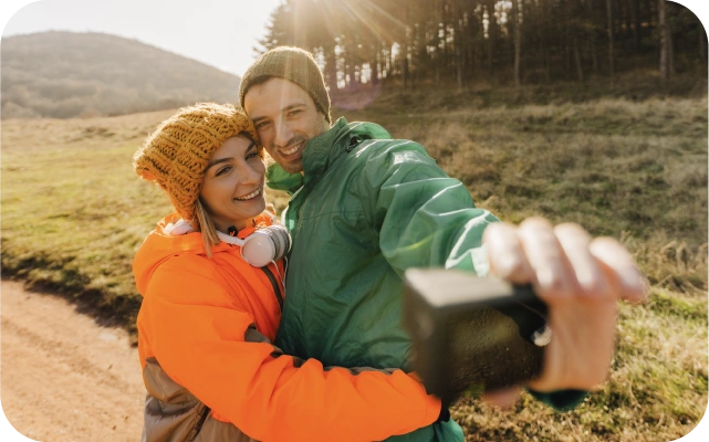 Vīrietis un sieviete stāv ārā dabā apķēruši viens otru; vīrietis tur kameru, lai uzņemtu pašbildi. Abi ir ģērbušies siltās, krāsainās jakās un cepurēs, fonā kalnains ainavas reljefs ar zāli un kokiem, saules stari spīd cauri koku rindai.