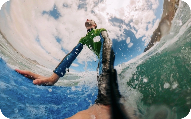 Vīrietis uz sērfa dēļa, fotografēta no zema leņķa ūdenī, ar viļņiem un ūdens šļakatām priekšplānā, fonā mākoņainas debesis un piekrastes klintis.