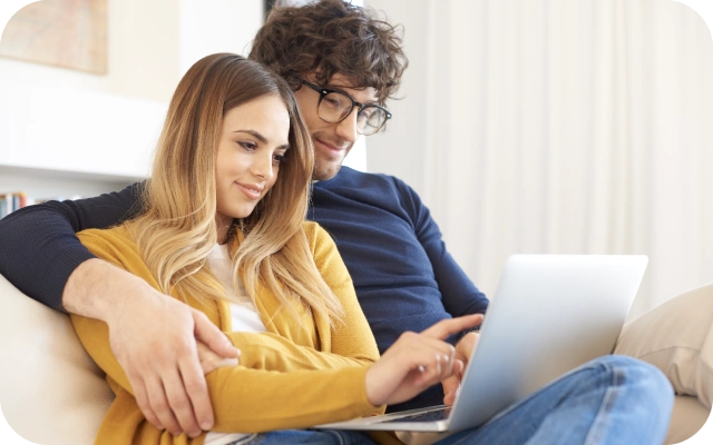 Uz dīvāna sēž vīrietis un sieviete, vīrietis ir apķēris sievieti ap pleciem, abi skatās portatīvajā datorā, kas ir sievietei klēpī.