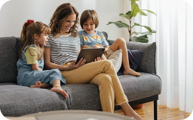Sieviete ar diviem bērniem sēž uz pelēka dīvāna un skatās planšetdatoru; fonā gaišā istaba ar augiem un aizkariem.