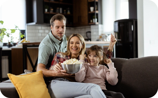 Ģimene sēž uz dīvāna viesistabā, sieviete ar vīrieti tur bļodu ar popkornu un skatās televizoru.