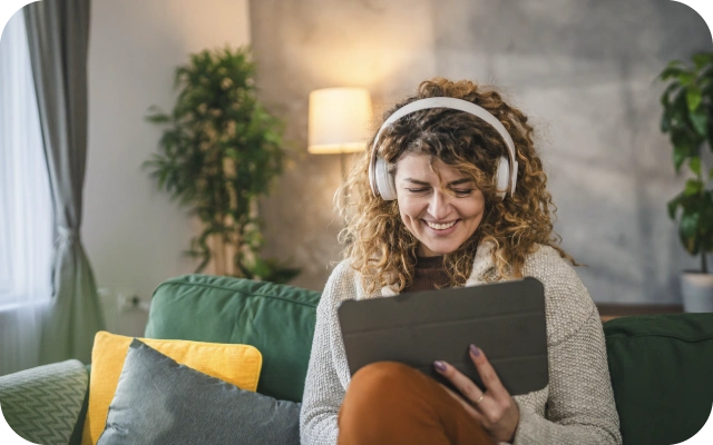 Sieviete ar cirtainiem matiem un austiņām sēž uz zaļa dīvāna ar spilveniem, turot rokās planšetdatoru.