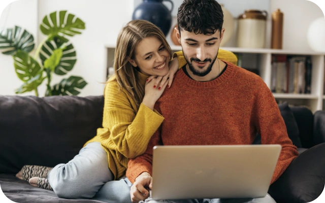 Sieviete un vīrietis sēž uz tumša dīvāna un skatās portatīvajā datorā, fonā zaļš istabas augs un grāmatu plaukts.