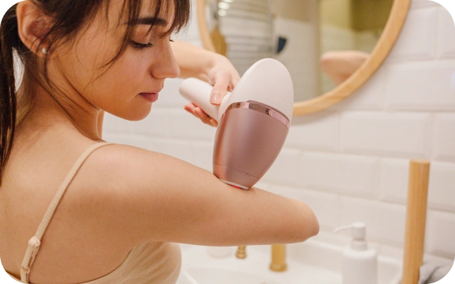 Sieviete vannasistabā izmanto IPL fotoepilatoru matiņu noņemšanai, virzot to gar rokas ādu. Fonā izlietne, spogulis un vannasistabas piederumi.