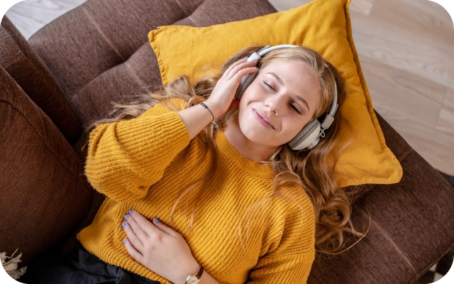 Sieviete gaišiem matiem un dzeltenā džemperī atpūšas, guļot uz dīvāna ar galvu uz dzeltena spilvena un klausoties mūziku austiņās.