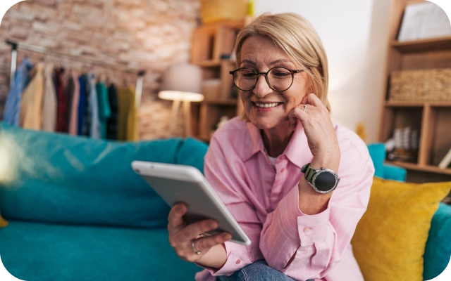 Sieviete sēž uz dīvāna mājīgā istabā, tērpusies rozā kreklā, un skatās planšetē. Fonā pakaramais ar apģērbiem, lampa un interjera priekšmeti.